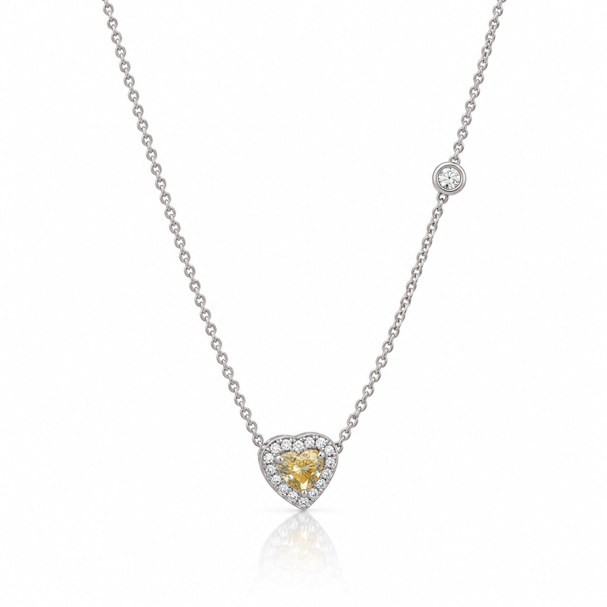 Silver-tone necklace with a yellow-centered heart pendant, a clear halo and an offset round station accent. Shown on a white background with a subtle reflection.